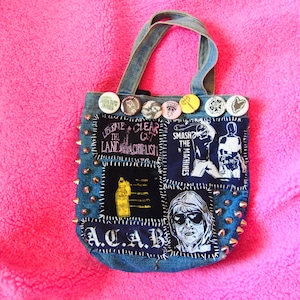 Pode incluir: Uma sacola de ganga com patches e tachas de metal. Os patches incluem texto e ilustrações. A sacola tem alças de ganga e é decorada com vários botões de alfinete. A sacola está em uma superfície rosa.