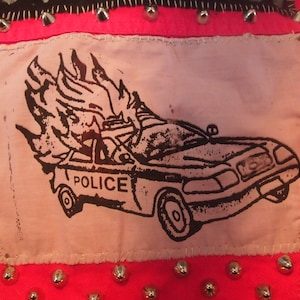 May include: A pink fabric patch with a black and white illustration of a police car on fire. The car has the word "POLICE" printed on the side.