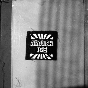 May include: A monochrome patch featuring the text "ABOLISH ICE" in white, bold lettering. The patch is rectangular with a sunburst design above and below the text. The image is in shades of grey.
