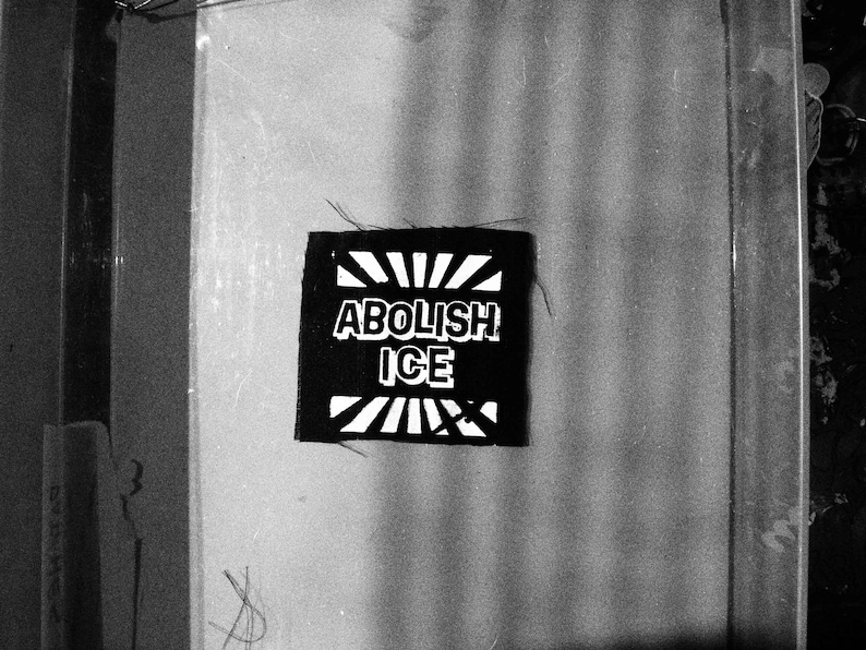 May include: A monochrome patch featuring the text "ABOLISH ICE" in white, bold lettering. The patch is rectangular with a sunburst design above and below the text. The image is in shades of grey.