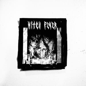 Puede incluir: Parche blanco y negro con el texto "Witch Fever" en una fuente en negrita y estilizada. El parche presenta un gráfico de un telón de escenario con una figura sombría detrás.