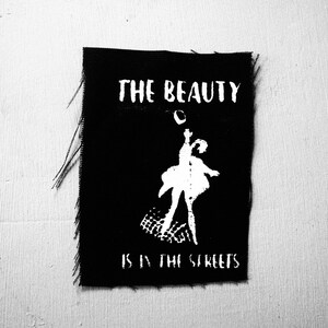 May include: Black fabric patch featuring white text and a ballerina illustration. The text states "THE BEAUTY IS IN THE STREETS". The ballerina is shown in a dynamic pose, with one arm raised and a flowing skirt.