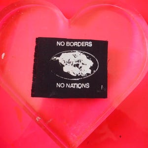 May include: A small, black fabric patch featuring the words "NO BORDERS" and "NO NATIONS" in white, with a white world map design in between. The patch is placed on a bright pink, heart-shaped surface.