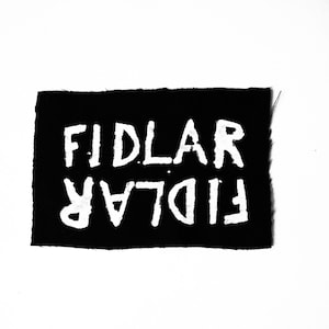 May include: A black rectangular patch with the word "FIDLAR" in large, white, distressed-style letters. Below the word is the same word, mirrored. The patch is set against a white background.