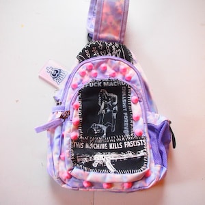 May include: A small, purple and pink tie-dye sling bag with a black patch featuring a graphic and text that reads "FUCK MACHO" and "THIS MACHINE KILLS FASCISTS". The bag has pink pom-poms and a small side pocket.