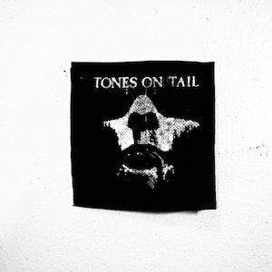 Puede incluir: Parche blanco y negro con el texto "TONES ON TAIL" en letras blancas. Una imagen estilizada de una persona con una prenda con capucha está en el centro del parche.