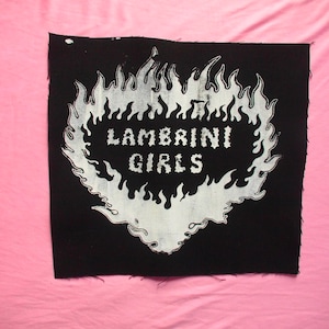 May include: Black fabric patch with the words "LAMBRINI GIRLS" in white, surrounded by a white flame design. The patch has a raw edge and is set against a pink background.