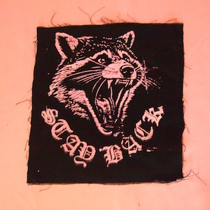 May include: Black fabric patch featuring a white image of a raccoon's face with an open mouth and sharp teeth. The text "Stay Back" is written in white below the image.