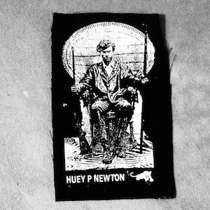 May include: Black and white image of Huey P. Newton sitting in a chair with a gun in his hand. The image is on a black patch with white text that reads "Huey P. Newton".