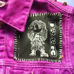 Puede incluir: Chaqueta vaquera morada vibrante con un parche negro con una figura de dibujos animados blanca y la palabra "Damage". La chaqueta tiene costuras blancas, tachuelas metálicas y un cuello clásico. La tela tiene un efecto tie-dye.