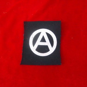 Può includere: Un cerotto di stoffa nero con un cerchio bianco che contiene un simbolo di anarchia, una lettera A stilizzata all'interno di un cerchio.