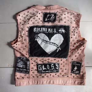 Puede incluir: Un chaleco vaquero rosa claro adornado con tachuelas plateadas y parches. Los parches presentan texto y gráficos, incluyendo "BIKINI KILL" con un corazón, "GL05.9" y "THE BEAUTY IS IN THE STREETS".
