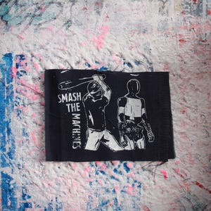 May include: Black rectangular patch with white graphic of a baseball player swinging a bat at a robot. Text on the patch reads "SMASH THE MACHINES". The patch is on a textured, multi-colored background.