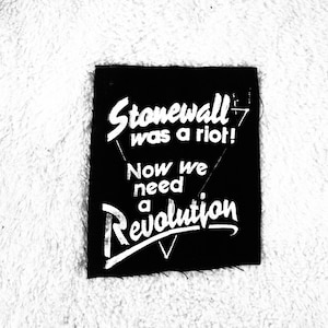 May include: Black and white patch with the text "Stonewall was a riot! Now we need a revolution" in white lettering.