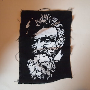 May include: Black and white patch with a stencil-like image of a man with a beard and mustache. The image is on a black fabric background.