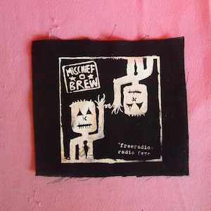 Puede incluir: Parche de tela negro con texto blanco e ilustraciones. El texto dice "Mischief Brew" y "freeradio radio fare". Las ilustraciones son de dos figuras de palo con caras.