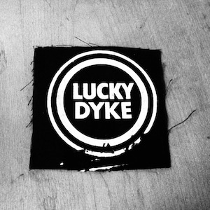 Peut inclure: Patch noir et blanc avec un cercle et le texte "LUCKY DYKE" à l'intérieur.