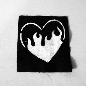 Pode incluir: Remendo de tecido preto com um desenho branco em forma de coração. O coração tem chamas saindo do topo.