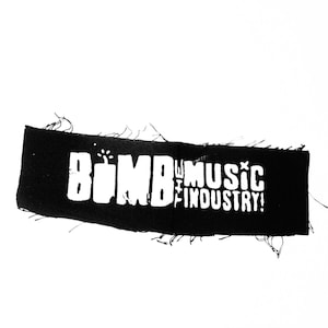 May include: Black rectangular patch with frayed edges. The patch features the white text "BOMB THE MUSIC INDUSTRY!" in a bold, stylized font. The design includes a small explosion graphic above the word "BOMB".
