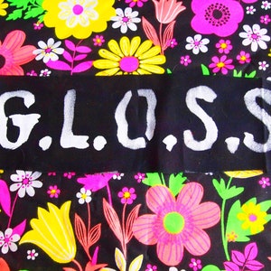 Pode incluir: Um patch de tecido preto com a palavra "G.L.O.S.S." em branco, com letras em estilo stencil. O patch está sobre um fundo vibrante de tecido floral com flores rosa, amarelas e brancas em um campo preto.