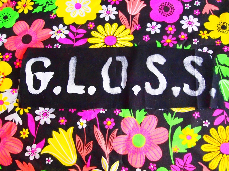 Peut inclure: Un patch de tissu noir avec le mot "G.L.O.S.S." en blanc, lettrage au pochoir. Le patch est pos&eacute; sur un fond de tissu floral vibrant avec des fleurs roses, jaunes et blanches sur un fond noir.