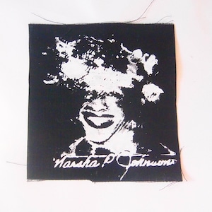 以下が含まれることがあります： マーシャ・P・ジョンソン、トランスジェンダー活動家の肖像画が描かれた白黒のアイロン接着パッチ。肖像画の下に「Marsha P. Johnson」の文字が書かれている。