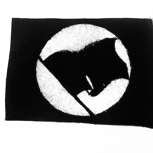 Puede incluir: Parche blanco y negro con un círculo blanco que contiene una bandera negra con un contorno blanco.