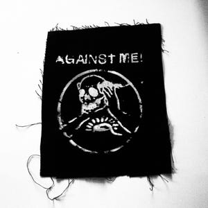Puede incluir: Parche blanco y negro con el texto "AGAINST ME!" y un gráfico de calavera.