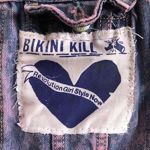 Puede incluir: Un parche de tela rosa cosido en una chaqueta vaquera. El parche presenta el texto "Bikini Kill" en letras negras, una forma de corazón en morado y el texto "Revolution Girl Style Now" en una pancarta morada.