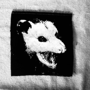 May include: Black and white patch featuring a detailed illustration of an opossum's head. The opossum is depicted with its mouth open, showcasing its teeth. The patch is square-shaped and likely made of fabric.