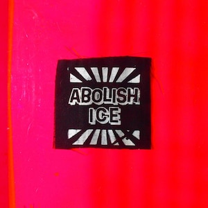 May include: A black patch with the words "ABOLISH ICE" in white block letters. The patch is set against a vibrant pink and red background.