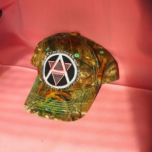 May include: Camouflage baseball cap with a circular patch featuring a pink letter "A" inside a black triangle and a white circle. The cap has green stitching and a curved brim. The hat is designed for outdoor activities.