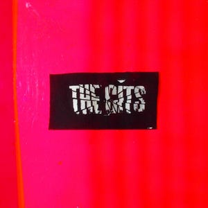 May include: A close-up of a vibrant image featuring a black rectangular patch with the text "THE KITS" in white, set against a bright pink and red background. The text appears in a distressed, stylized font.