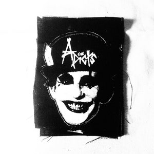 May include: Black and white iron-on patch featuring a person wearing a hat with the text "The Adicts" on it.