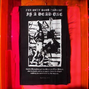 以下が含まれることがあります： 「THE ONLY GOOD FASCIST IS A DEAD ONE」のテキストが入った白黒のグラフィックパッチ。画像には、逆さまに吊るされた2つの人物が描かれています。テキストにはベニート・ムッソリーニとクララ・ペタッキについて書かれています。1995年4月28日の日付も含まれています。