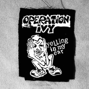 May include: Black and white patch featuring the text "OPERATION IVY" above a cartoon figure covering their ears. The text "yelling in my ear" is also present. The patch is rectangular.