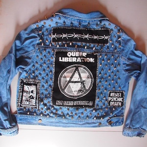 May include: Light blue denim jacket adorned with silver studs and patches. Patches include barbed wire, the words "QUEER LIBERATION" and "NOT STATE SANCTIONED", and the phrase "RESIST PSYCHIC DEATH". A unique punk rock style.