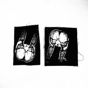 Könnte beinhalten: Zwei schwarze Stoffaufnäher mit weißen Illustrationen. Einer zeigt eine Rückansicht einer sich bückenden Person, der andere eine ähnliche Illustration aus einem anderen Winkel. Die Aufnäher sind quadratisch und haben einen rohen Rand.