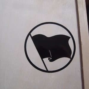 Puede incluir: Un círculo blanco y negro con una bandera negra en su interior. La bandera ondea hacia la derecha y tiene una línea diagonal que la atraviesa.