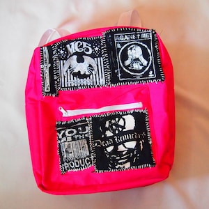 Op de afbeelding: Een felroze rugzak met verschillende zwart-witte patches die erop zijn genaaid. De patches hebben tekst, waaronder "Against Me!" en "You Are The Product".