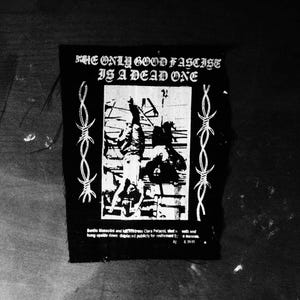 Op de afbeelding: Zwart-witte patch met de tekst "THE ONLY GOOD FASCIST IS A DEAD ONE". De patch toont een afbeelding van ondersteboven hangende figuren, omlijst door prikkeldraad. Extra tekst is onderaan aanwezig.