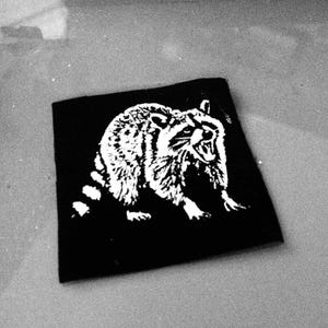 Könnte beinhalten: Ein schwarzer quadratischer Aufnäher mit einer weißen Illustration eines Waschbären. Der Waschbär mit offenem Maul und gestreiftem Schwanz vor schwarzem Hintergrund.