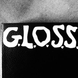 May include: Close-up of a black leather surface with the word "GLOSS" written in white, stylized lettering.