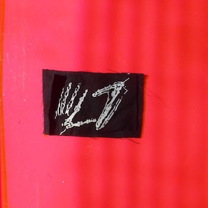 Puede incluir: Un parche rectangular negro con texto blanco sobre un fondo rosa brillante. El texto blanco es un logotipo o diseño estilizado. El parche está cosido sobre la superficie rosa, posiblemente como una etiqueta o emblema.