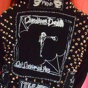 May include: Black jacket with silver cone studs and white stitched detailing. The back features a large patch with the words "Christian Death" and an illustration. Additional text includes "Fiend" and "Only Theatre of Pain."