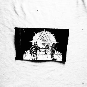 Pode incluir: Patch preto e branco com um gráfico de três figuras caminhando em direção a uma estrutura triangular com as letras "L" e "F". O patch é retangular com uma borda irregular.