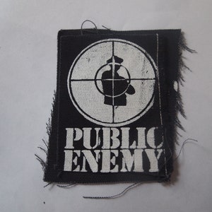 May include: Black and white embroidered patch with a white circle containing a stylised silhouette of a person in a target sight. The text "PUBLIC ENEMY" is below the circle.
