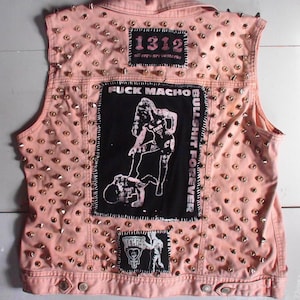 May include: A pink denim vest covered in silver metal studs. The back features patches with the text "1312 all cops are bastards", "FUCK MACHO BULLSHIT FOREVER", and a graphic of a person. A punk rock style.
