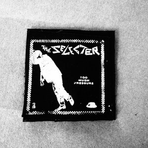 Op de afbeelding: Zwart-witte patch met de tekst "The Selector" en "Too Much Pressure" in witte letters. In het midden van de patch is een silhouet van een persoon afgebeeld.