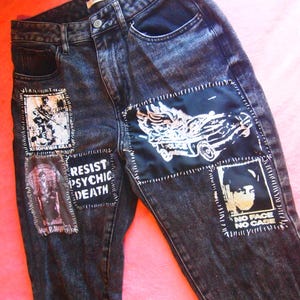May include: Black denim jeans with multiple patches sewn on. The patches feature black and white images and text, including "Resist Psychic Death" and "No Face No Case".